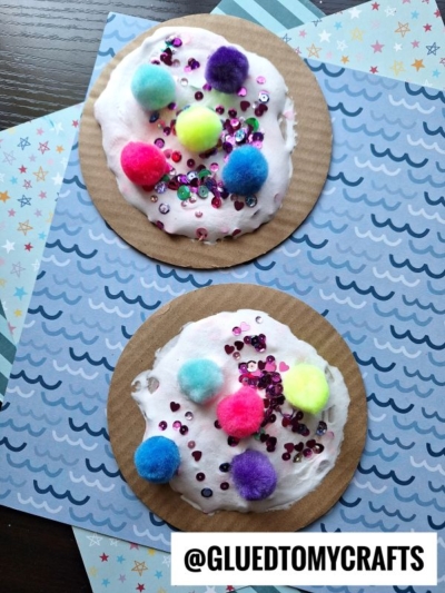 Two round cardboard circles decorated with white puffy paint, colorful pom-poms, and sequins to look like cookies—a fun puffy paint crafts idea! They're displayed on blue patterned paper. @GLUEDTOMYCRAFTS is written at the bottom.