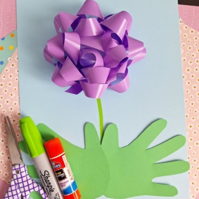 A craft project for a 3D flower card for mom, featuring a purple gift bow as a flower on blue paper, with green handprint cutouts as leaves and stem. Scissors, markers, and a glue stick are nearby. Patterned papers decorate the background.