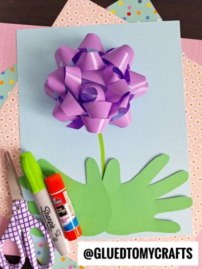 A craft project with a purple gift bow as a flower, green paper handprints as leaves, and a drawn stem on light blue paper. Scissors, glue stick, markers, and patterned papers are shown nearby.