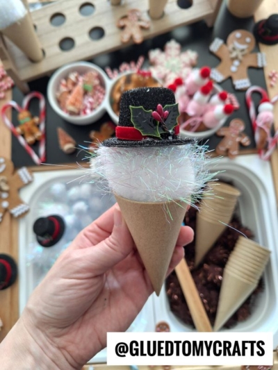A hand holds a craft cone decorated as a snowman’s head with a black hat, red ribbon, holly, and sparkly white material—perfect for pom pom ice cream play. Festive craft supplies and treats are visible in the background.