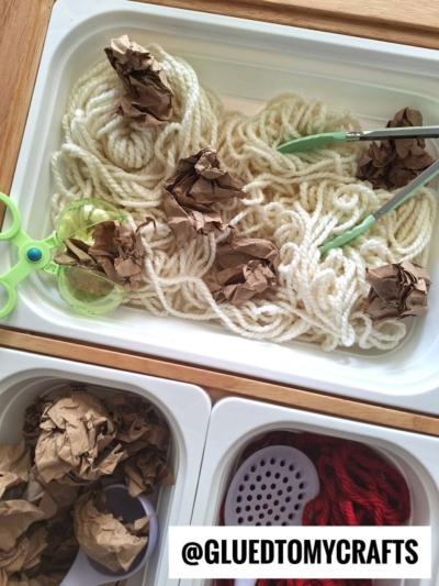 A sensory bin filled with white yarn, brown crumpled paper, green scissors, and green tongs. Nearby bins hold more brown paper, a white scoop, and red yarn. @GLUEDTOMYCRAFTS is written at the bottom.