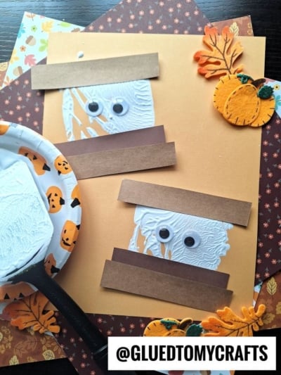 Two mummy crafts made from white paint, googly eyes, and brown paper strips are displayed on an orange background with fall-themed decorations, including leaves and pumpkins. A spatula stamped s'mores craft adds to the festive scene. A plate with white paint is nearby.