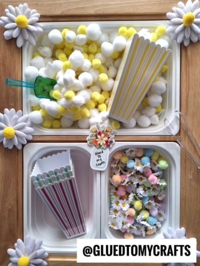 A sensory bin activity with containers of yellow and white pom-poms, pastel pom-poms, plastic scoops, paper popcorn boxes, artificial daisies, and a sign reading “Glued To My Crafts.”.