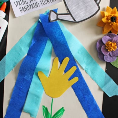 A spring craft project featuring a yellow paper handprint flower, blue crepe paper as rain, a watering can cutout, felt flowers, glue, and a sign reading Spring Showers Bring Handprint Flowers. Text reads Free Printable!!!.
