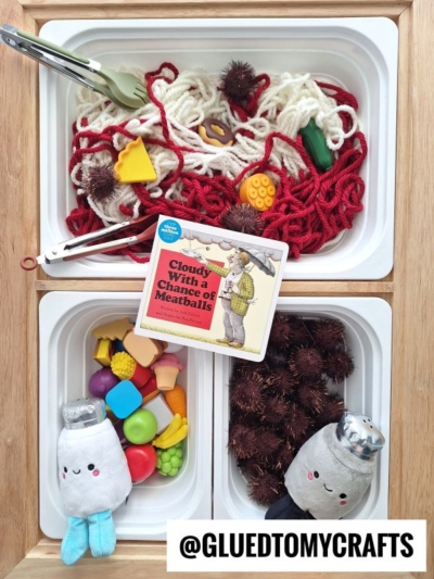 A sensory bin features craft yarn spaghetti, toy food items, brown pom-poms, a Cloudy With a Chance of Meatballs book, plush dolls dressed as salt and pepper shakers, and a variety of colorful plastic food for imaginative play.