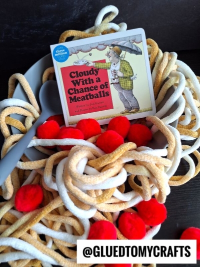 A bowl with tan and white yarn resembling spaghetti, red pom-poms as meatballs, a gray fork, and a Cloudy with a Chance of Meatballs book, with @GLUEDTOMYCRAFTS at the bottom.