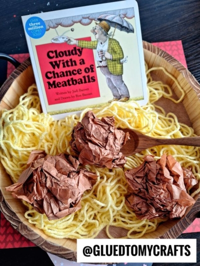 A wooden bowl filled with yellow yarn and brown crumpled paper sits on a table, resembling spaghetti and meatballs. Behind the bowl is the book Cloudy With a Chance of Meatballs. The text @GLUEDTOMYCRAFTS is at the bottom.