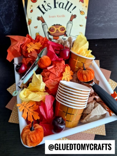 A tray with orange and gold cups, fake pumpkins, red apples, paper leaves, crumpled tissue paper, scissors, and the children’s book Its Fall! displayed in the background—inspired by a tissue paper ice cream sensory bin activity.