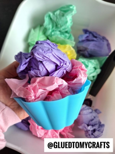A hand holds a blue silicone cupcake liner filled with crumpled pink and purple tissue paper, resembling a tissue paper ice cream sensory bin, with more colorful tissue paper balls in a white container below. Text reads @GLUEDTOMYCRAFTS.