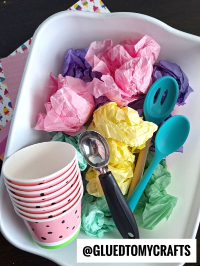 A white tray holds colorful crumpled tissue paper, stacked watermelon-themed paper cups, a metal ice cream scoop, and blue plastic serving spoons. A label reads @GLUEDTOMYCRAFTS.