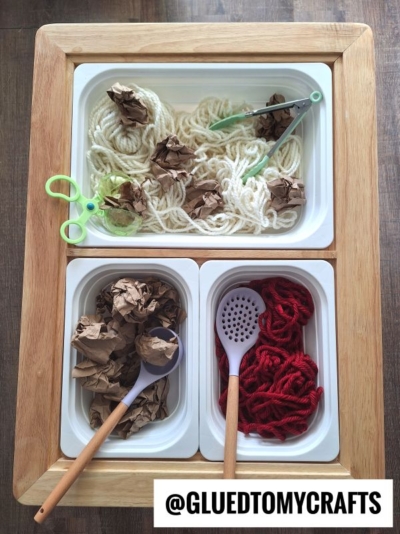Three bins with play materials: top bin has white yarn, brown paper, and green play tweezers; left bin has crumpled brown paper and a large spoon; right bin has red yarn and a slotted spoon. @GluedToMyCrafts label at bottom.