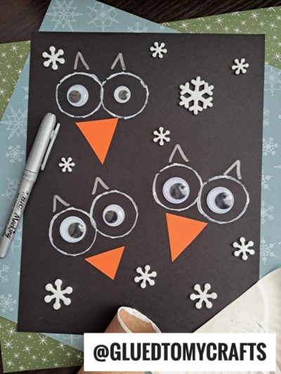 A black paper features three owl faces made with googly eyes, orange paper beaks, and white marker outlines. White snowflakes are scattered around. A silver marker and craft supplies are nearby. Text reads @GLUETOMYCRAFTS.