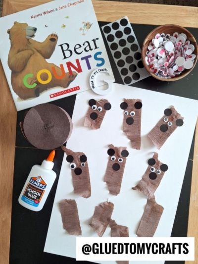 A children’s craft setup inspired by the book Bear Counts features torn crepe paper craft ideas—brown paper for bear faces on white sheets, with googly eyes, black noses, glue, and supplies around. The handle @gluedtomycrafts is visible.