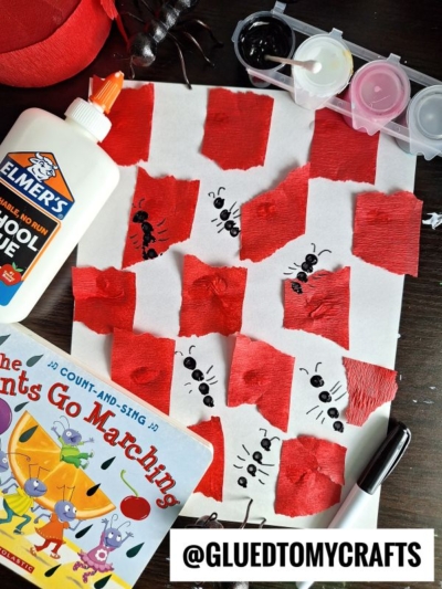 A children's craft scene with torn red tissue squares glued to white paper, black marker ant faces, a glue bottle, paint palette, black marker, and The Ants Go Marching book—perfect for INSECT CRAFT IDEAS. Labeled @GLUEDTOMYCRAFTS.