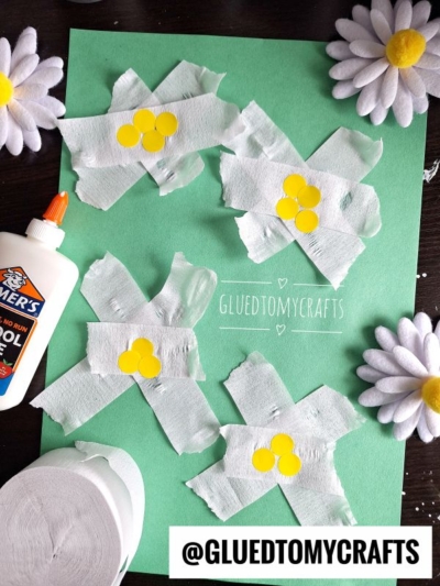 A green paper with four flowers made from white tissue and yellow circles, resembling daisies. Glue, tissue roll, and artificial daisies are nearby—perfect inspiration for crepe paper craft ideas. The watermark @GLUEDTOMYCRAFTS is at the bottom.
