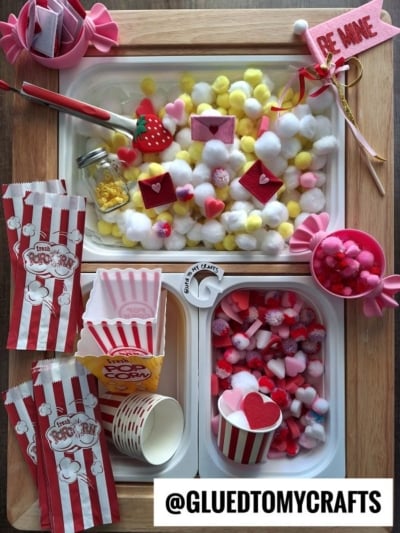 A craft tray filled with colorful pompoms, heart shapes, paper popcorn bags, small cups, beads, and Valentine-themed decorations, including a “Be Mine” wand. The display has a cute and festive Valentine’s Day theme.