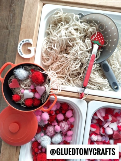 A sensory bin setup for craft yarn spaghetti play with yarn noodles, red and pink pom-poms, heart shapes, a toy pot, and kitchen utensils. The @GLUEDTOMYCRAFTS handle is labeled at the bottom.