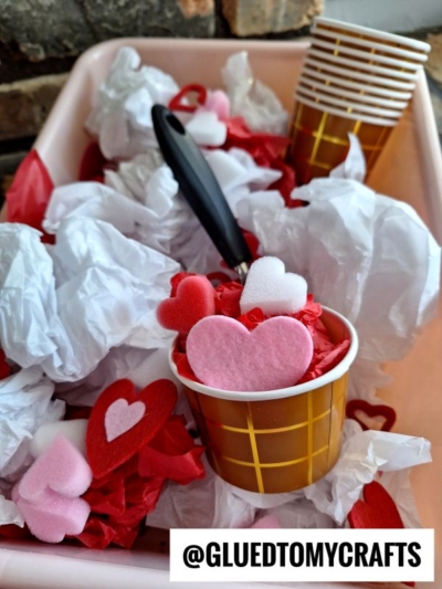Small brown cups filled with red, pink, and white felt hearts sit among crumpled white tissue paper in a tray, resembling a tissue paper ice cream sensory bin. A black spoon rests in one cup. The image is marked with @GLUEDTOMYCRAFTS.