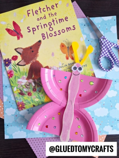 A children’s book titled “Fletcher and the Springtime Blossoms” sits on polka dot paper next to purple scissors and a pink popsicle stick butterfly craft decorated with sequins, googly eyes, and a wooden stick body.