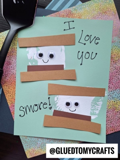 A handmade card with two marshmallow characters made of paper, googly eyes, and brown strips for smores, with the text I love you smore! on a pastel background. A spatula stamped smores craft and colorful papers are nearby.