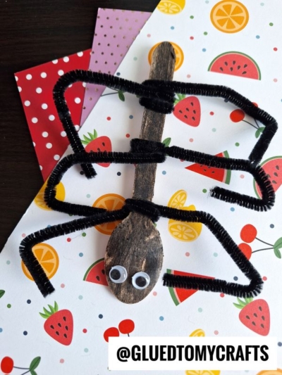 A wooden spoon painted black with googly eyes and black pipe cleaners as spider legs is displayed on colorful fruit-themed paper, making it perfect for INSECT CRAFT IDEAS. @GLUEDTOMYCRAFTS text appears at the bottom.