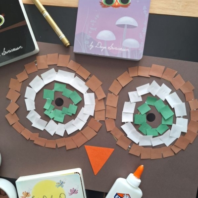 A mosaic owl craft made with brown, white, and green paper squares on black paper, surrounded by a glue bottle, gold pen, and owl-themed books. The handle @GLUEDTOMYCRAFTS is at the bottom.