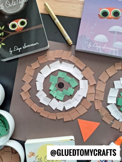 A mosaic craft resembling an owls face made from cut paper squares in brown, green, and white, displayed on a brown background. Owl-themed books, a pen, and craft supplies surround the artwork.