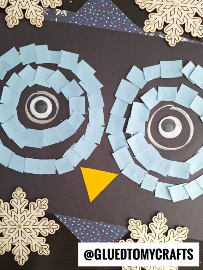 A craft owl face made with blue paper squares for eyes, large googly eyes in the center, and a yellow triangle beak, on black paper, decorated with snowflake cutouts. @GLUEDTOMYCRAFTS is written at the bottom.