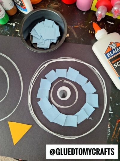 Black paper with a white spiral outline, small blue paper squares glued along the spiral, a yellow triangle in the corner, a bowl of blue squares, white glue, and bottles of craft paint. Text: @GLUEDTOMYCRAFTS.