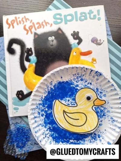 Children’s book "Splish, Splash, Splat!" features a black cat and yellow duck in water; up front, a bubble wrap stamped duck craft on blue-painted paper. Beads and paper are nearby. @GLUEDTOMYCRAFTS.