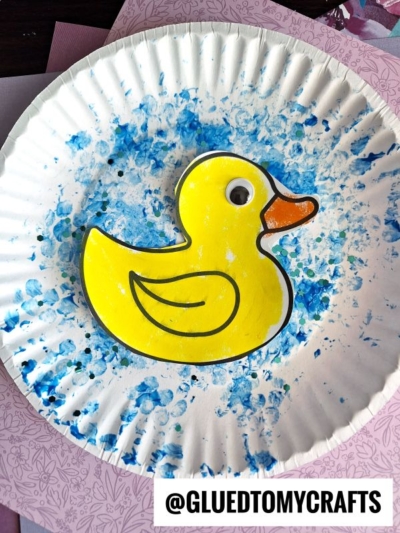A white paper plate decorated with blue paint splatters features a bubble wrap stamped duck with a googly eye and an orange beak glued at the center. The image includes the text @GLUEDTOMYCRAFTS at the bottom.