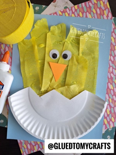 A handmade chick craft made with yellow crepe paper strips, googly eyes, an orange paper beak, and a paper plate half for the body on a blue background—perfect for those seeking fun crepe paper craft ideas, with craft supplies beside it.
