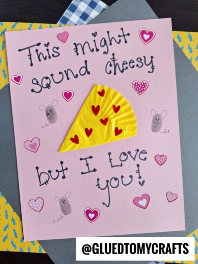 A handmade cupcake liner card craft featuring the words This might sound cheesy but I love you! Decorated with a yellow cheese slice, red hearts, and tiny gray mice. The Instagram handle @GLUEDTOMYCRAFTS is at the bottom.