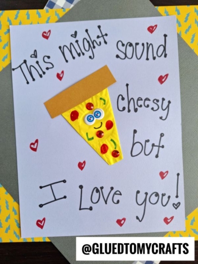 A handmade cupcake liner card features a smiling pizza slice with googly eyes and the message This might sound cheesy but I love you! written in playful lettering, surrounded by small red hearts. Perfect for fun card crafts lovers.