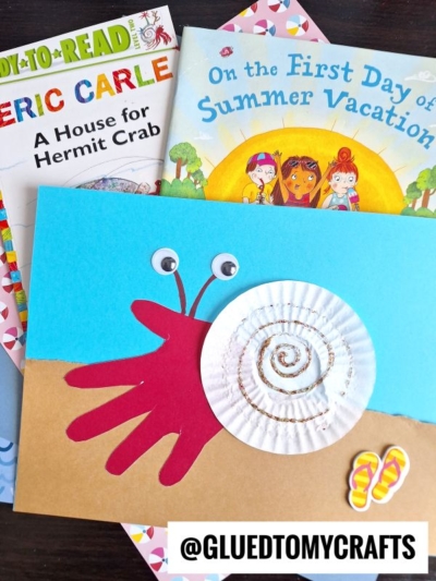 A children’s craft features a handprint red crab with googly eyes and a white cupcake liner shell, placed over two colorful childrens books about summer and hermit crabs, with a sticker of flip-flops nearby.