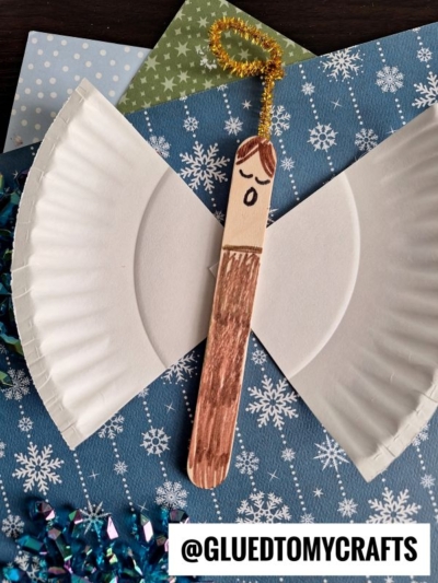 A Christmas angel craft made from a popsicle stick with a drawn face, brown hair, and robe, a gold pipe cleaner halo, and wings crafted from a cut white paper plate, displayed on blue snowflake paper.