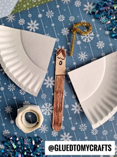 A craft project on a snowflake-patterned surface features a paper plate cut into two wings, a popsicle stick angel with a drawn face and brown hair, gold pipe cleaner halo, glue dots, and blue sequins. Text reads @GLUEDTOMYCRAFTS.