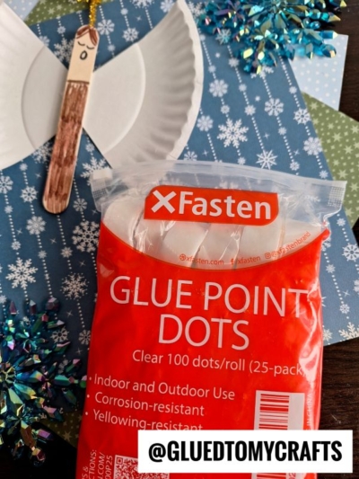 A package of XFasten Glue Point Dots sits on a blue snowflake-patterned surface next to a popsicle stick angel craft and a white plate cut in half to form wings. The handle @GLUEDTOMYCRAFTS is displayed.