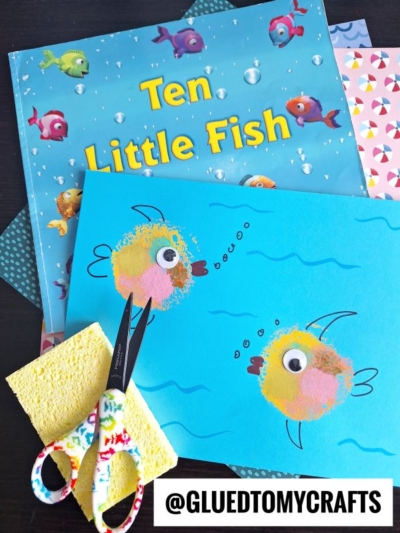 A childrens book titled Ten Little Fish lies beside a blue paper craft featuring a sponge painted fish, bubbles, drawn waves, a sponge, and patterned scissors. @GLUEDTOMYCRAFTS is written at the bottom.