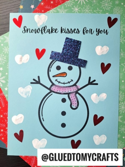 A handmade snowman card on blue paper with glittery blue hat, pink scarf, heart decorations, and the text "Fingerprint snowman kisses for you." The Instagram handle @GLUEDTOMYCRAFTS appears at the bottom.