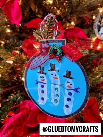A handmade Christmas ornament shaped like a bauble features three fingerprint snowman designs, each decorated with hats, scarves, and gems, hanging on a decorated tree with red ribbon and holiday lights.