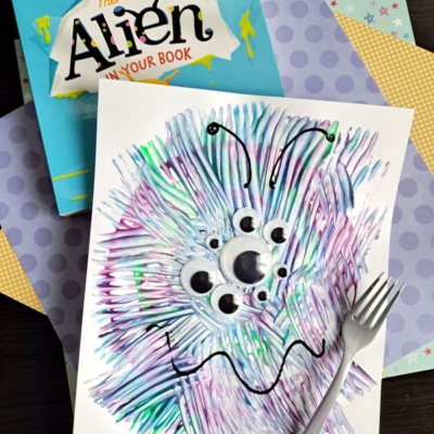 A children’s book titled “There’s an Alien in Your Book” sits next to a painted alien craft with googly eyes, created using a plastic fork, displayed on colorful polka-dot and patterned papers.