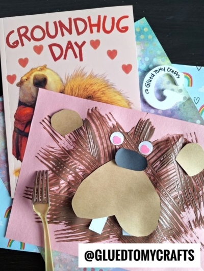 A children’s book titled Groundhug Day lies next to a pink craft paper featuring a fork painted groundhog, crafted from brown cut-out shapes. A plastic fork used for painting and a sticker reads @GLUEDTOMYCRAFTS.