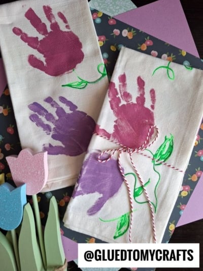 Two white cloths decorated with pink and purple handprint flowers make charming Mother’s Day keepsakes. One is tied with red and white string. Colorful paper and wooden tulip decorations are in the background.