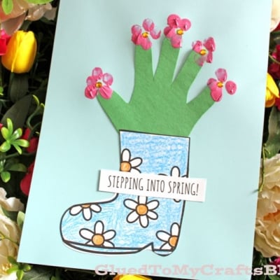 A handmade card shows a blue rain boot with drawn flowers and a green handprint sprouting pink flower stickers, with a sign reading STEPPING INTO SPRING! surrounded by colorful real flowers.