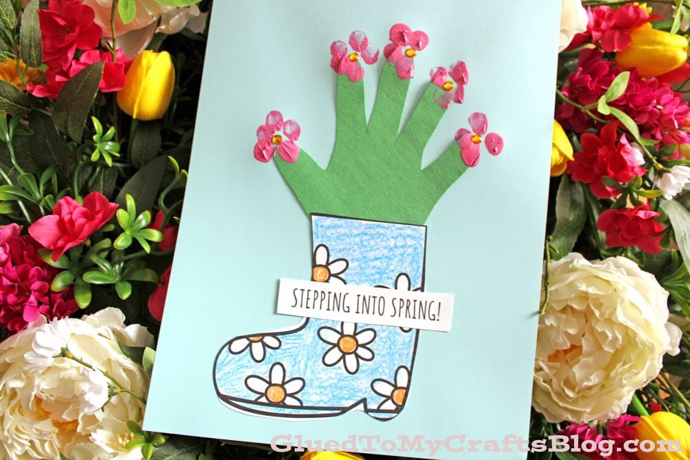 A handmade card shows a blue rain boot with drawn flowers and a green handprint sprouting pink flower stickers, with a sign reading STEPPING INTO SPRING! surrounded by colorful real flowers.