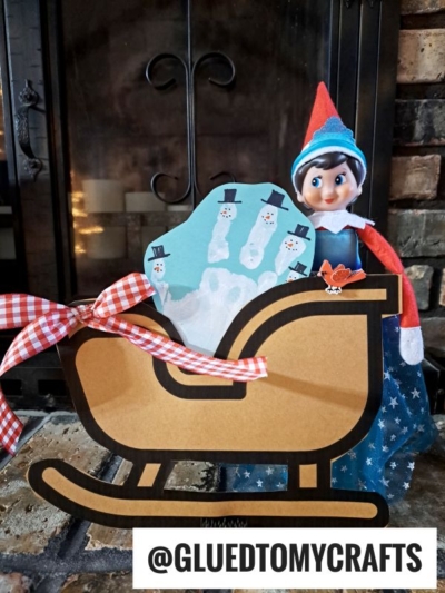 An Elf on the Shelf doll stands next to a Christmas sleigh keepsake, decorated with a child’s handprint painted as snowmen and tied with a red and white gingham bow. A fireplace glows warmly in the background.