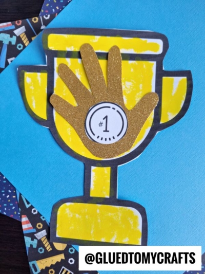 A handmade Handprint Trophy card features a yellow base, gold glitter hand, and #1 medal at the center, set against a blue background with patterned paper. The text @GLUEDTOMYCRAFTS appears at the bottom.