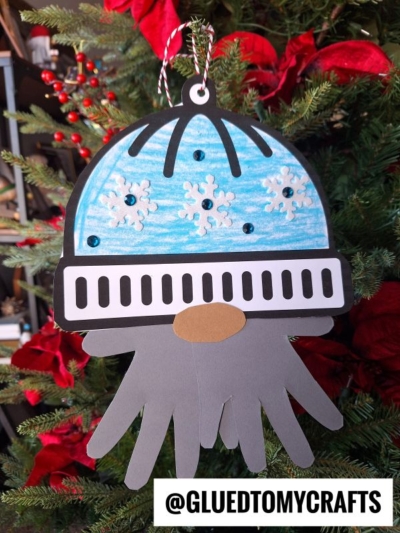 A handmade winter gnome keepsake ornament features a blue, snowflake-adorned hat and gray handprint tentacles, hanging on a Christmas tree with red poinsettias. Text at the bottom reads @GLUEDTOMYCRAFTS.