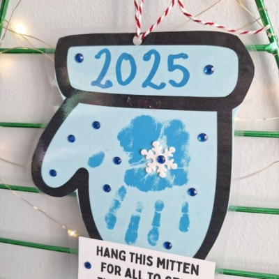 A handmade mitten height ornament labeled 2025 features a blue handprint, a white snowflake in the center, and blue gems. It hangs by red-and-white string with a note below and @GLUEDTOMYCRAFTS at the bottom.